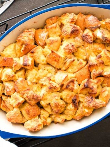 A person places a blue oval baking dish filled with golden-brown bread pudding on a cooling rack. Two forks rest on a nearby plate, and the person is holding the dish with white cloths.