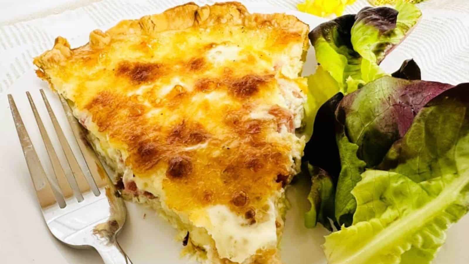 Slice of Quiche Lorraine on white plate next to fork and green salad.