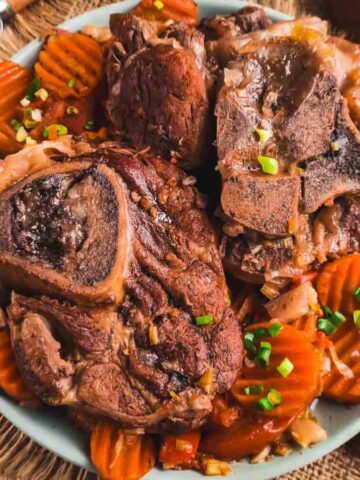 A plate of cooked lamb chops sits on a bed of sliced, cooked carrots. The dish is garnished with chopped green onions. Utensils, a wooden spoon with spices, and a small bowl of more green onions are nearby on a burlap cloth surface.