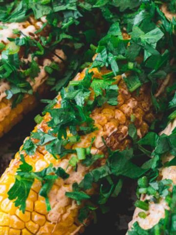 Grilled corn on the cob covered with a creamy sauce and topped with chopped green herbs, arranged closely together on a surface.