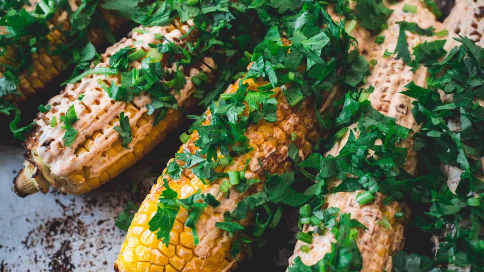 Grilled corn on the cob covered with a creamy sauce and topped with chopped green herbs, arranged closely together on a surface.