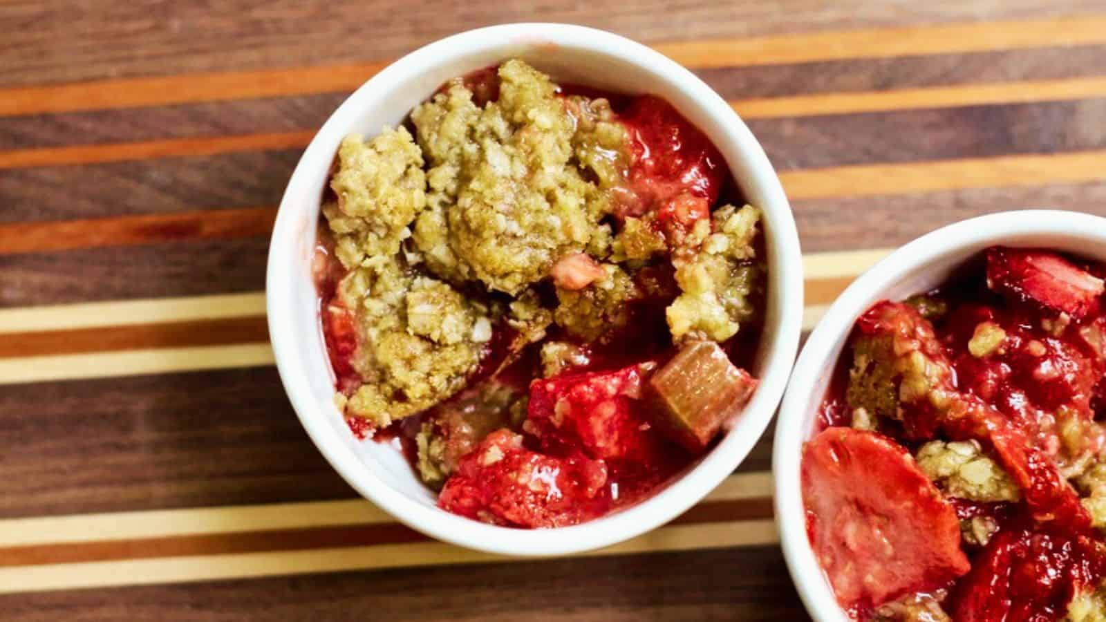 Two white ramekins filled with strawberry rhubarb crisp, featuring a golden-brown crumbly topping and vibrant red fruit filling, placed on a striped wooden surface. This fresh and fruity dessert is perfect for a summer vacation treat.