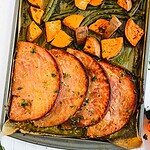 Slices of cooked ham steak are arranged on a sheet pan with roasted sweet potato chunks and green beans. The dish is sprinkled with herbs and brown sugar, appearing freshly baked with a delightful browning on the edges.