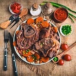 A plate of Osso Buco with bone, garnished with sliced carrots and green onions. Surrounding the dish are cutlery, spice containers, diced tomatoes, cherry tomatoes, and green chilies on a burlap surface for that slow-cooked charm.