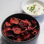 A black bowl filled with crispy Air Fryer Beet Chips sits on a white surface. Behind it, a white bowl holds a creamy dip topped with chopped green herbs.