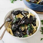 A white bowl filled with creamy penne pasta and garlic mussels, garnished with chopped parsley, sits on a marble surface. Two utensils rest in the bowl. A blue pot with more mussels is partially visible in the background.