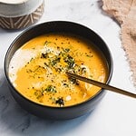 A black bowl filled with creamy orange carrot soup, garnished with herbs, ground pepper, and a drizzle of cream. A gold spoon rests inside the bowl, on a light marble surface next to a tan cloth—an inviting slow cooker recipe.