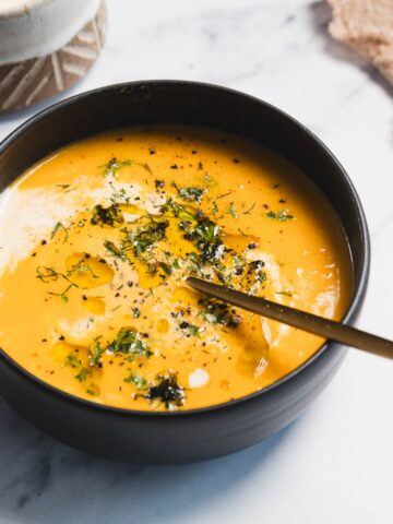 A black bowl filled with creamy orange carrot soup, garnished with herbs, ground pepper, and a drizzle of cream. A gold spoon rests inside the bowl, on a light marble surface next to a tan cloth—an inviting slow cooker recipe.