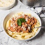 A white bowl filled with penne pasta, topped with a chunky tomato meat sauce, grated cheese, and a fresh basil leaf. A spoon rests in the bowl, and a towel and cheese dish are nearby on a marble surface.