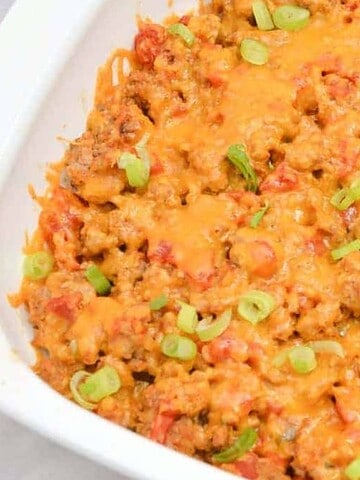 A white baking dish filled with a cheesy casserole topped with melted cheddar cheese and sliced green onions. The casserole appears to contain ground meat and diced tomatoes.