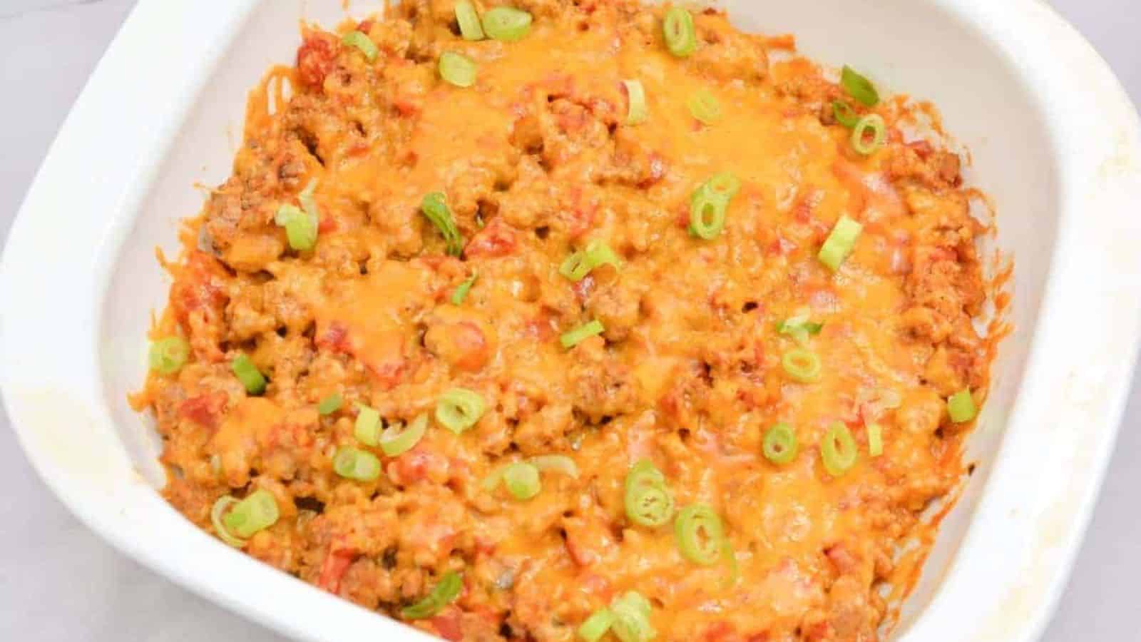 A white baking dish filled with a cheesy casserole topped with melted cheddar cheese and sliced green onions. The casserole appears to contain ground meat and diced tomatoes.