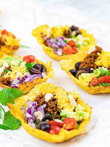 Eight mini taco bowls filled with ground meat, shredded cheese, lettuce, tomatoes, olives, and red cabbage are arranged on a white tray, with fresh cilantro sprigs placed beside them.