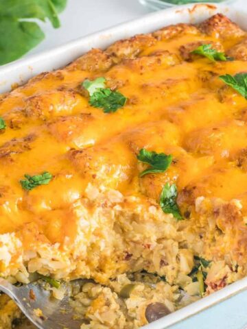 A rectangular casserole dish filled with cheesy enchiladas topped with melted cheddar cheese and garnished with parsley. A portion is missing, exposing the filling, with a serving spoon resting in the dish. Fresh spinach leaves are in the background.