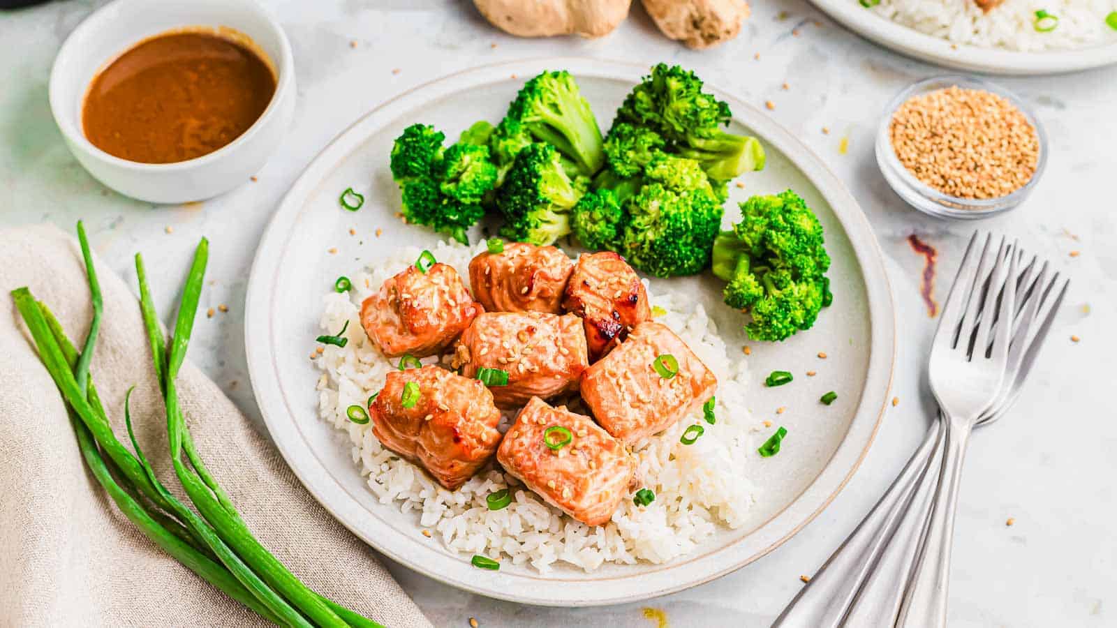 A plate with grilled salmon cubes on white rice, topped with chopped green onions, accompanied by broccoli florets. Ideal for air fryer enthusiasts, a bowl of sauce, sesame seeds, and fresh green onions are nearby. Silver forks rest on the table.