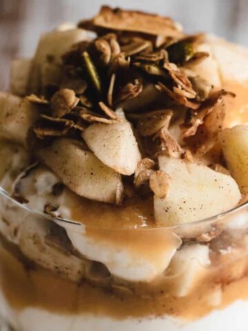 A glass filled with layers of yogurt, caramel sauce, diced apples, and granola, placed on a white surface with a wooden background.