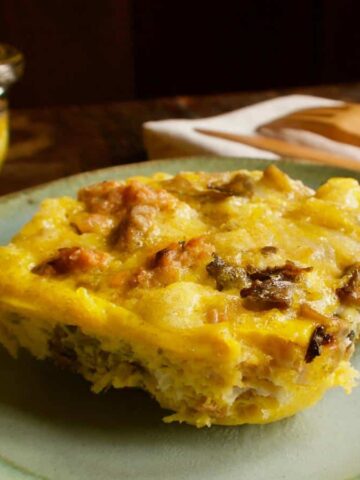 A slice of sausage egg bake is placed on a greenish-blue plate. The dish includes visible chunks of sausage and other ingredients. In the background, a larger portion of the bake is in a glass dish. A wooden fork and knife are nearby.