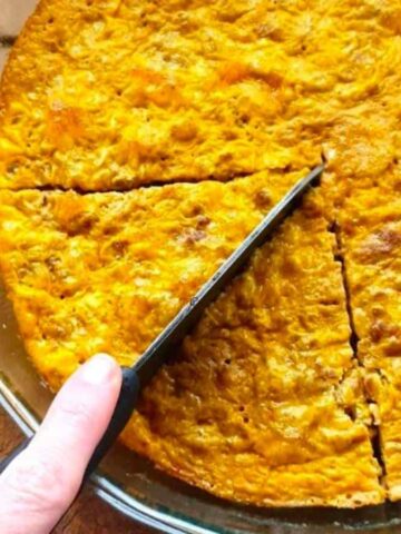 A hand holds a knife cutting slices from a round baked dish with a golden-brown top, placed in a clear glass pie dish on a wooden surface. Three forks are visible nearby.