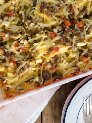 A rectangular baking dish filled with a baked breakfast casserole containing ground beef, shredded cheese, hash browns, and chopped red peppers. A slice is served on a white plate with a blue rim, accompanied by a fork.