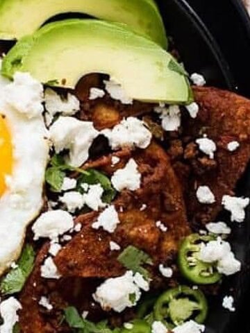 A plate of chilaquiles topped with a sunny-side-up egg, sliced avocado, radishes, jalapeño, crumbled cheese, and chopped cilantro, served on a black dish.