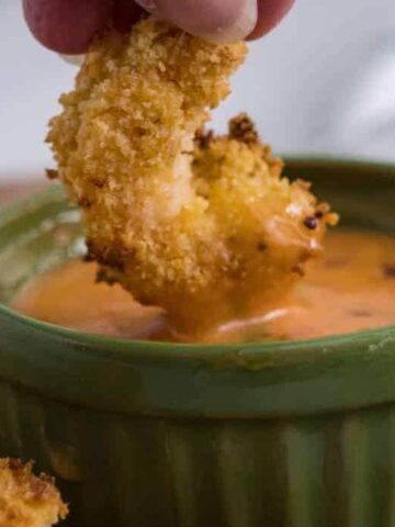 A hand dips a piece of breaded, crispy food into orange-colored sauce in a green ceramic bowl. A checkered cloth is visible in the blurred background.