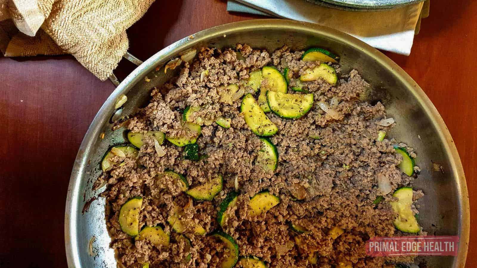 A stainless steel pan on a wooden surface contains cooked ground beef, sliced zucchini, and diced onions, seasoned with black pepper&mdash;perfect for budget meals. A beige cloth and a pale napkin are partially visible nearby.