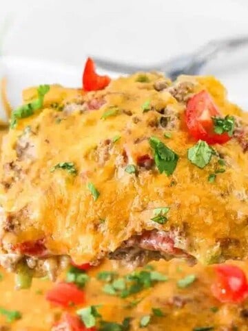 A serving of taco casserole is being lifted with a metal spatula from a baking dish. The casserole is topped with melted cheese, diced tomatoes, and chopped herbs. A jar and some greens are visible in the background.