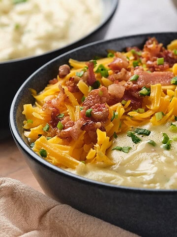 A close-up of a bowl of creamy soup topped with shredded cheddar cheese, chopped bacon, and chopped chives. Another bowl of soup is partially visible in the background.