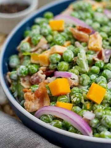 A bowl filled with a salad made of green peas, diced cheddar cheese, red onion slices, crispy bacon pieces, and a creamy dressing. The salad is served in a dark-rimmed bowl on a woven placemat.