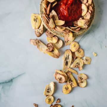 A bowl of red salsa partially surrounded by dried banana chips, with several chips scattered on a light gray marble surface.