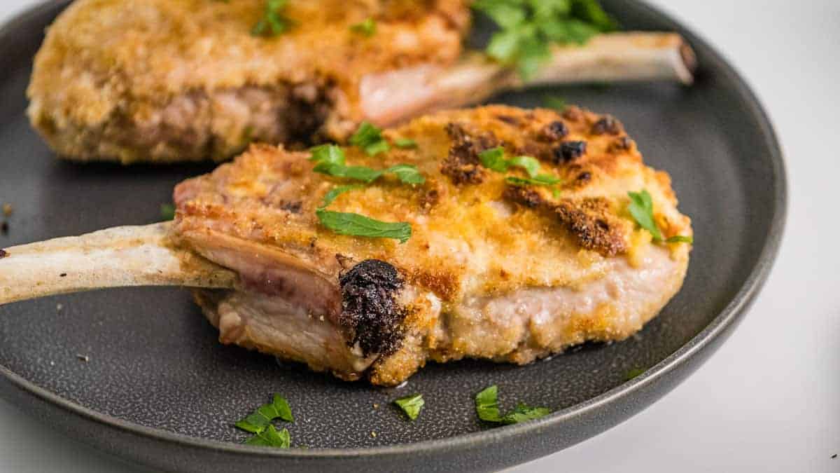 Close-up of a breaded pork chop with a bone, served on a sleek black plate. The chop, cooked to perfection in the air fryer, is garnished with fresh parsley. Another crispy-chopped masterpiece is partially visible in the background, highlighting the texture of its golden coating.