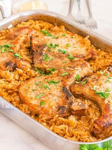 A rectangular roasting pan contains four cooked pork chops on a bed of rice, garnished with chopped parsley. The dish is on a marble countertop, with a folded white cloth and a small plate of mixed greens nearby.
