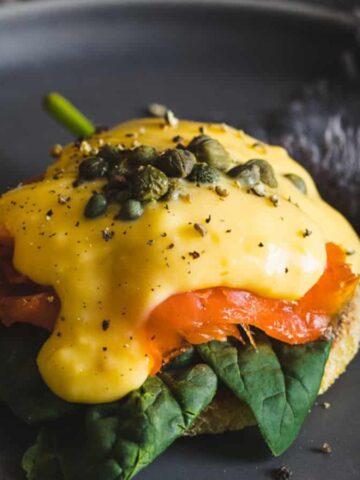 A plate contains a dish of eggs Benedict, featuring spinach, smoked salmon, and capers on an English muffin, topped with yellow hollandaise sauce. A fork is placed on the right side of the plate with a gray napkin nearby.
