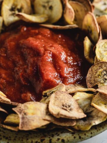 A speckled bowl filled with tomato sauce in the center, surrounded by a ring of crispy, thinly sliced plantain chips on a light surface.