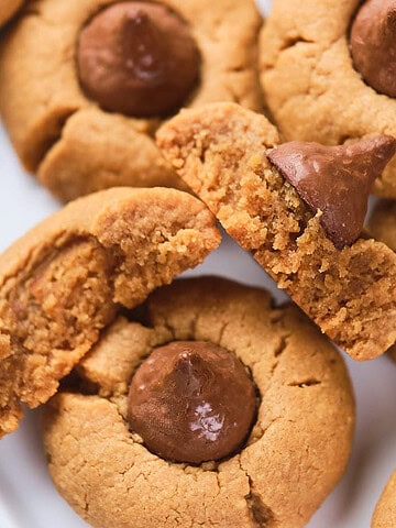 A close-up of several peanut butter cookies topped with chocolate kisses, some whole and one broken in half, arranged on a white plate.