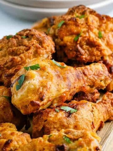 A wooden board topped with several pieces of golden-brown fried chicken, garnished with small green herb leaves. A salt or pepper shaker and stack of plates are visible in the background.