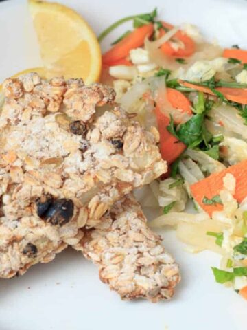 A white plate with two pieces of oat and raisin crusted baked chicken, a serving of sautéed cabbage with carrot slices, and a lemon wedge garnish.