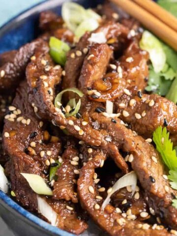 A blue bowl filled with sliced cooked beef topped with sesame seeds and green onions, served with cucumber slices, fresh cilantro, and chopsticks resting on the edge.