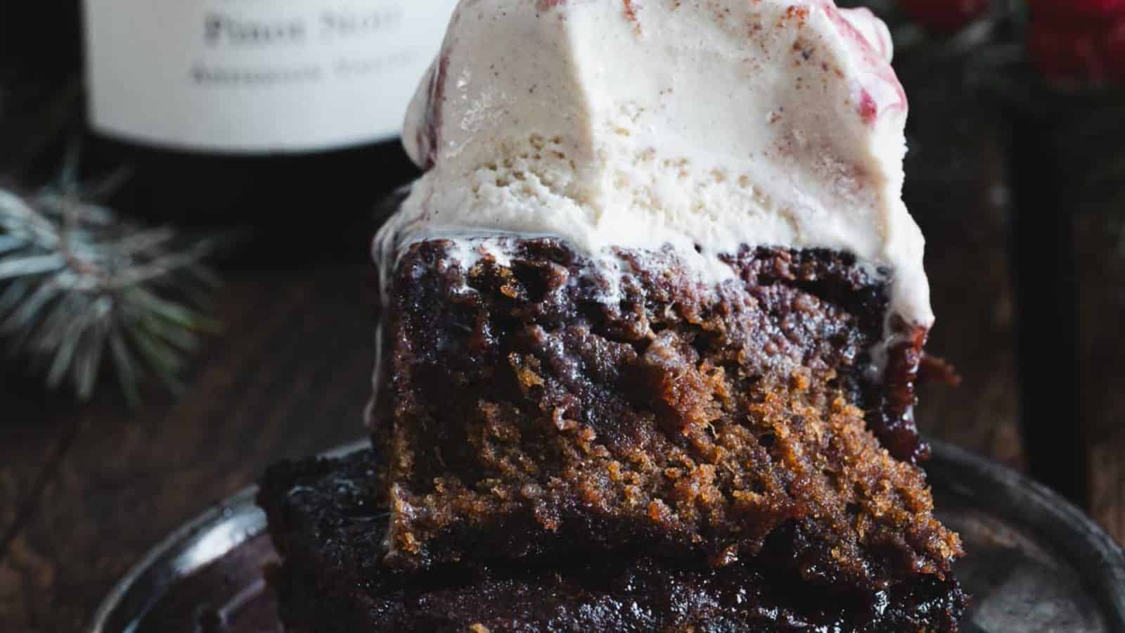 A slice of dark cake with a scoop of vanilla ice cream on top, drizzled with berry sauce, sits on a plate. Red berries and evergreen branches are in the background on a wooden surface.