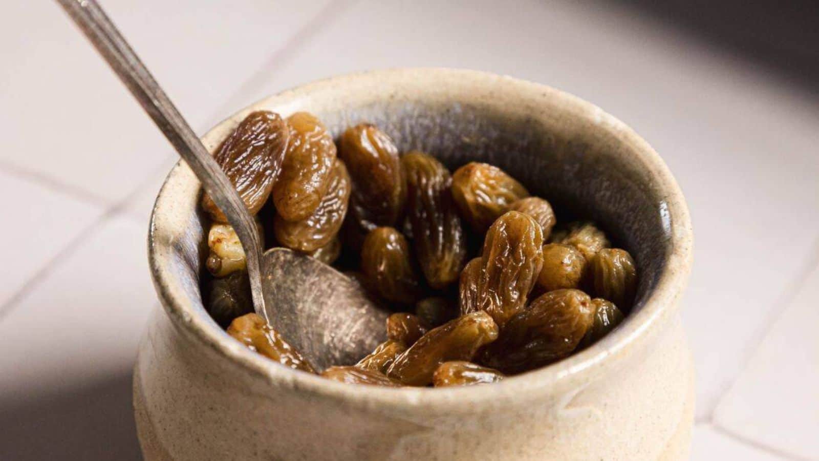 A ceramic bowl filled with golden raisins, with a metal spoon resting inside.
