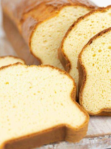 A loaf of bread with a golden-brown crust is partially sliced on a wooden cutting board. Four slices are visible, and the bread has a light, airy texture.