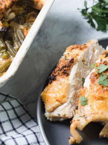 A plate with pieces of roasted chicken breast topped with herbs and a side of mixed vegetables sits next to a baking dish containing more chicken and vegetables. A checkered towel and sprigs of parsley are nearby.