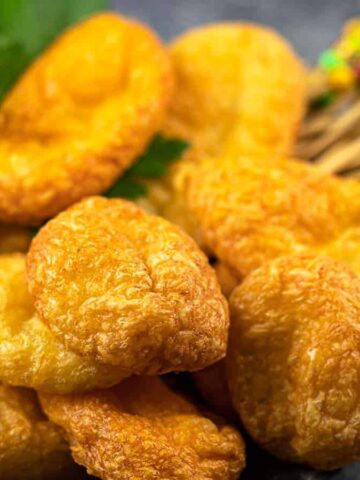 A close-up of several puffed and golden-brown fried snacks stacked on a dark surface, with colorful toothpicks and a green garnish in the background.