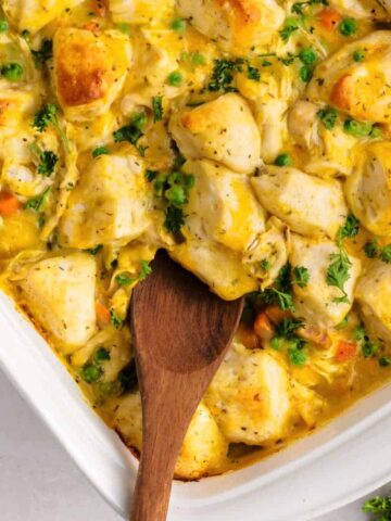 A wooden spoon rests in a white baking dish filled with a casserole of biscuit pieces, chicken, peas, carrots, and creamy sauce, garnished with chopped parsley. A striped towel and fresh parsley are nearby.