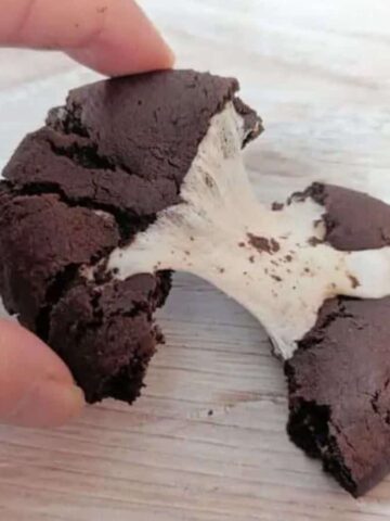 A hand pulls apart a chocolate cookie revealing a stretchy, melted marshmallow filling inside. The cookie is on a light-colored wooden surface.