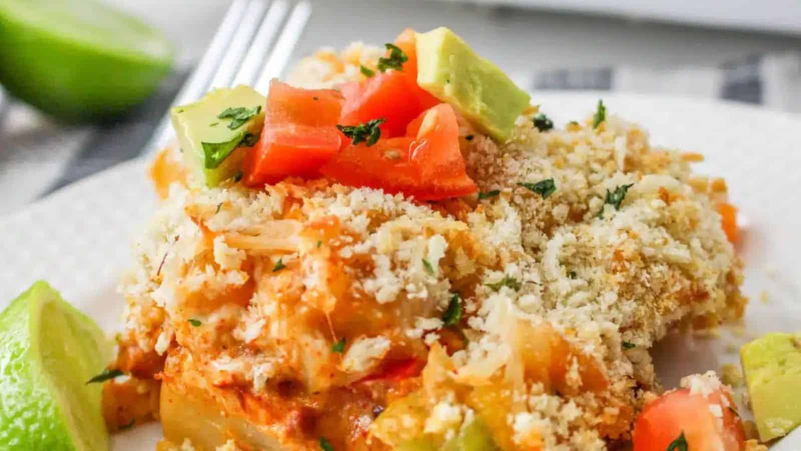 A serving of baked casserole with a crumbly topping on a plate, accompanied by guacamole and avocado slices. A casserole dish and more guacamole are in the background, placed on a wooden board.