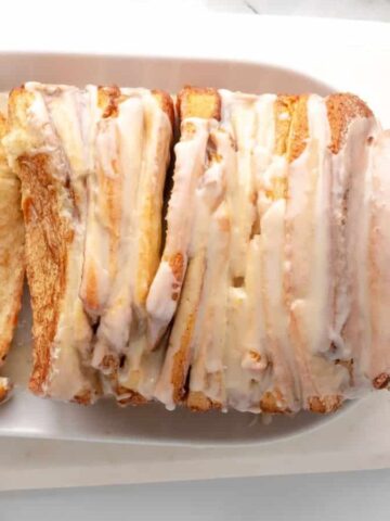 A loaf of pull-apart bread covered with white icing is displayed on a white serving board, with two slices partially separated. Cinnamon sticks and a spoon with icing are also visible nearby.