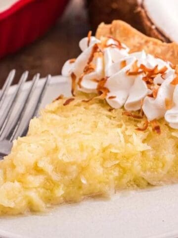 A slice of coconut cream pie topped with whipped cream and toasted coconut is on a white plate with a fork, set on a wooden surface.