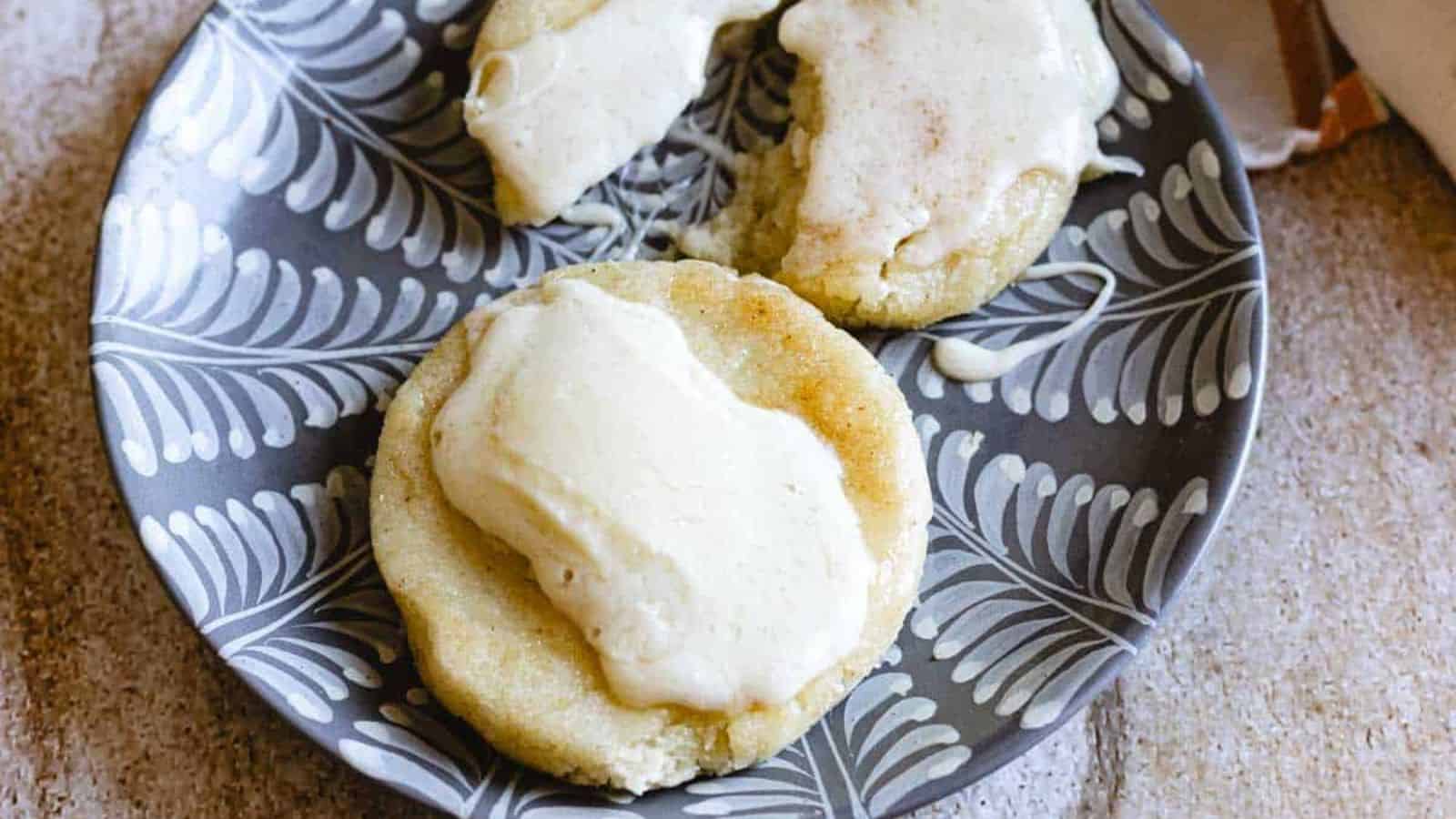 A patterned plate holds three round sugar cookies topped with a layer of white icing. One cookie is broken in half, showing a soft, slightly crumbly texture inside.