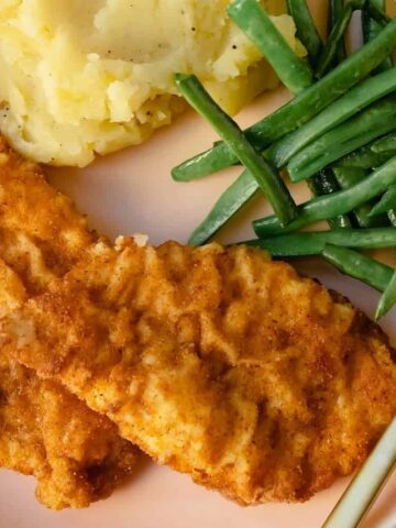 A plate with two breaded chicken tenders, mashed potatoes, and green beans. A gold fork is placed on the right side of the plate.