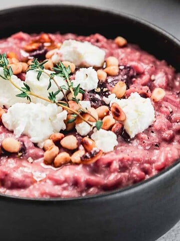 A black bowl filled with pink beetroot risotto, topped with dollops of white goat cheese, pine nuts, and a sprig of fresh thyme, placed on a light-colored surface.